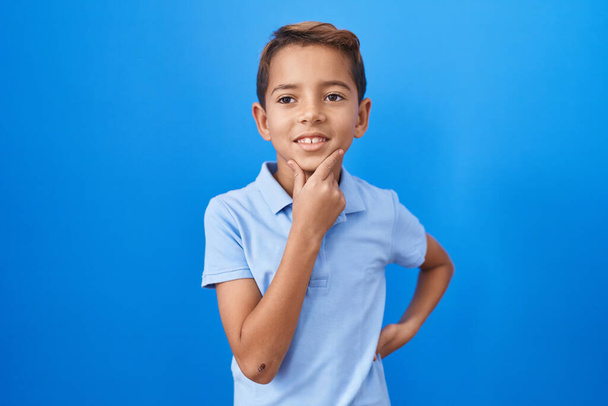 A kis spanyol fiú alkalmi kék pólóban mosolyog, magabiztosan néz a kamerába keresztbe tett karokkal és kézzel az állán. pozitív gondolkodás.  - Fotó, kép