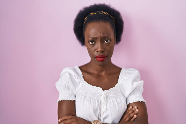 ピンクの背景の上に立っている巻き毛を持つアフリカの女性は懐疑的で緊張し、交差した腕で顔に不承認の表現。否定的な人.  - 写真・画像
