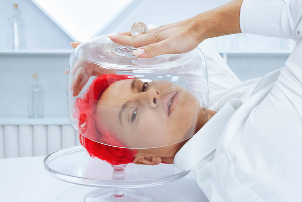 Жінка з коротким яскраво-червоним волоссям лежить на столі і покриває голову скляним куполом. концепція психічних проблем
. - Фото, зображення