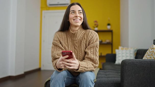 Young beautiful hispanic woman using smartphone sitting on sofa at home - 写真・画像
