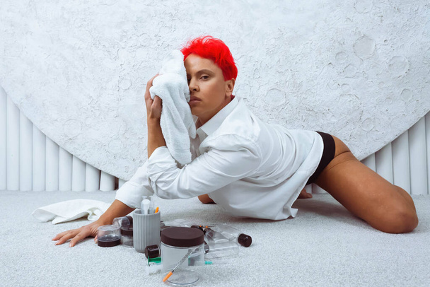 Kobieta z krótkimi, jasnoczerwonymi włosami siedząca na podłodze w białym pokoju, trzymająca słoiki z narkotykami. Koncepcja uzależnienia od narkotyków. - Zdjęcie, obraz