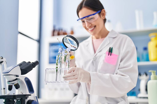 Jovem mulher vestindo uniforme cientista olhando flores com lupa no laboratório - Foto, Imagem