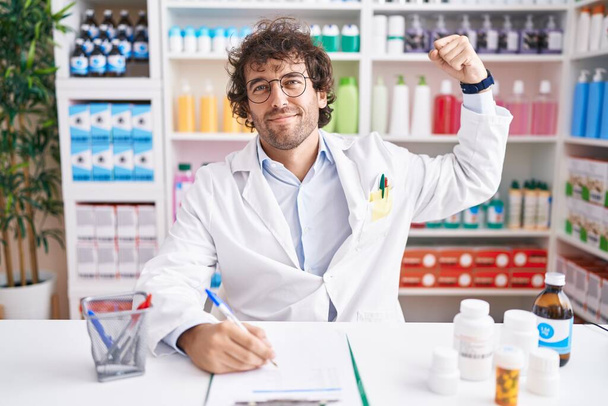 Hispanic young man working at pharmacy drugstore strong person showing arm muscle, confident and proud of power  - Photo, Image