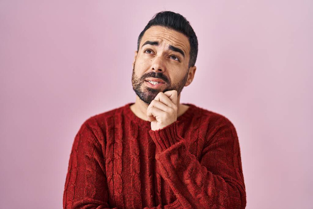 あごの質問について考える上で手でピンクの背景にカジュアルなセーターを身に着けてひげを持つ若いヒスパニック系の男性は、集中的な表現。思慮深い顔で笑顔。疑わしい概念.  - 写真・画像