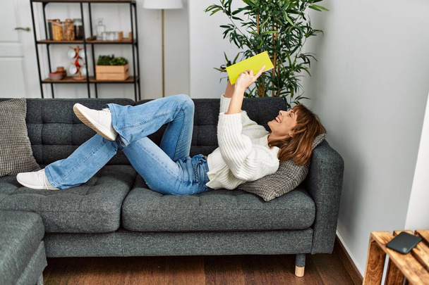 Молода біла жінка читає книгу, що лежить на дивані вдома
 - Фото, зображення