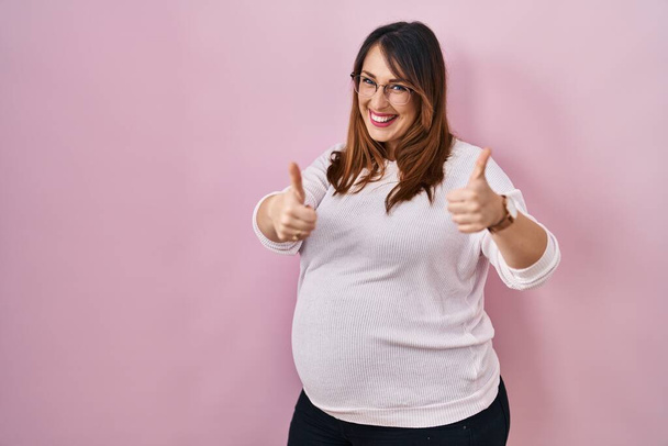 Беременная женщина, стоящая на розовом фоне и одобряющая позитивный жест рукой, улыбается и радуется успеху. жест победителя.  - Фото, изображение
