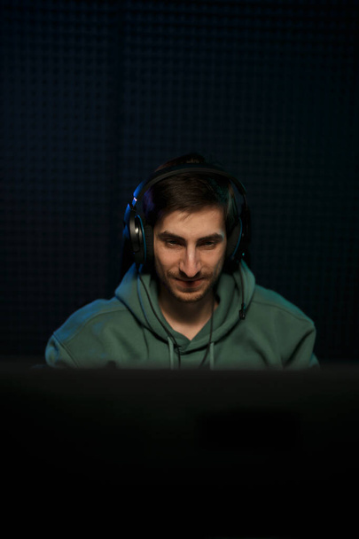 Podekscytowany młody mężczyzna cybersport gracz w słuchawki uśmiecha się szczęśliwie, ciesząc się zwycięstwem po udanej grze wideo na komputerze w ciemnym pokoju - Zdjęcie, obraz