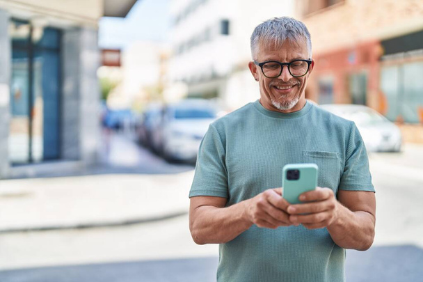 Middelbare leeftijd grijsharige man glimlachend vol vertrouwen met behulp van smartphone op straat - Foto, afbeelding