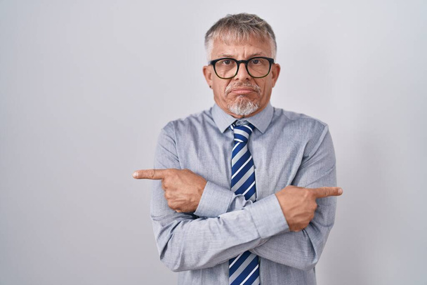 Hispanic business man with grey hair wearing glasses pointing to both sides with fingers, different direction disagree  - Foto, imagen