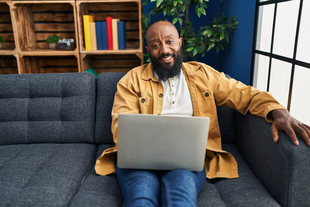 Африканський американець, який користується ноутбуком вдома сидячи на дивані, виглядає позитивним і щасливим, стоїть і посміхається з упевненою посмішкою.  - Фото, зображення