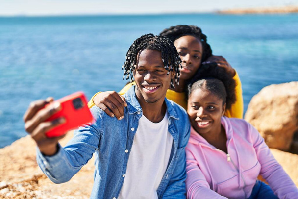 Afrykańscy amerykańscy przyjaciele robiący selfie przy smartfonie siedzącym na skale nad morzem - Zdjęcie, obraz