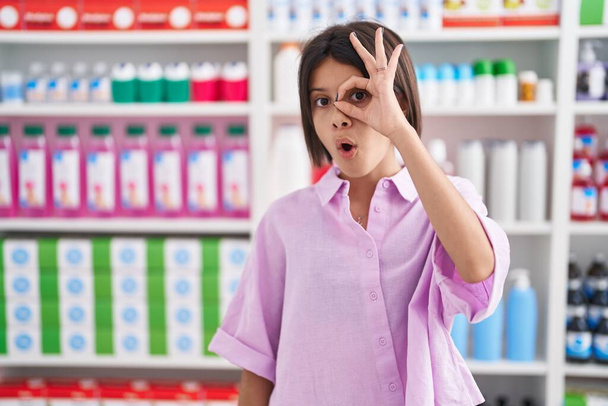 Jong meisje in de apotheek drogisterij doet ok gebaar geschokt met verrast gezicht, oog kijken door vingers. ongelovige uitdrukking.  - Foto, afbeelding
