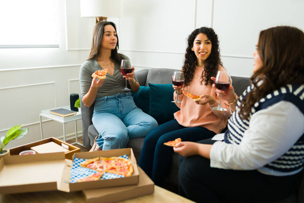 Glückliche Freundinnen lächeln, während sie Wein trinken und gemeinsam Pizza essen, während sie es sich im Wohnzimmer gemütlich machen - Foto, Bild