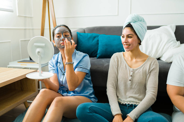 Aantrekkelijke beste vrienden zetten gezichtsmaskers en huidverzorgingsproducten op in hun pyjama terwijl ze ontspannen tijdens een leuk slaapfeestje - Foto, afbeelding