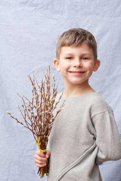 Portrait of handsome preschool boy with bouquet of spring flowers on blue background. child holding branches of flowering willow in hand, smiling. Happy easter. Palm Sunday. Springtime. - Valokuva, kuva