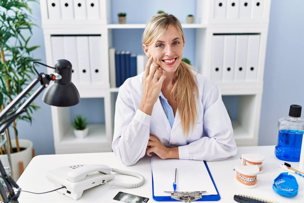 Young blonde woman wearing dentist uniform sitting on table at clinic - Fotó, kép