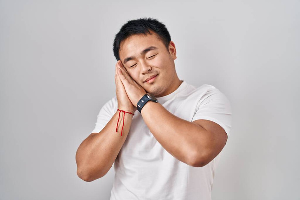 Młody Chińczyk stojący nad białym tłem śpiący zmęczony snem i pozujący razem dłońmi, uśmiechnięty zamkniętymi oczami.  - Zdjęcie, obraz