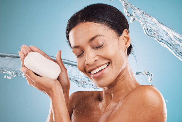 Νερό splash, χαρούμενη και μαύρη γυναίκα με σαπούνι για περιποίηση δέρματος, αυτοαγάπη ή αυτοφροντίδα στο studio σε μπλε φόντο. Χέρια, ντους και υγιή Αφρικής κορίτσι πλύσιμο, περιποίηση ή τον καθαρισμό του προσώπου ή το σώμα της. - Φωτογραφία, εικόνα