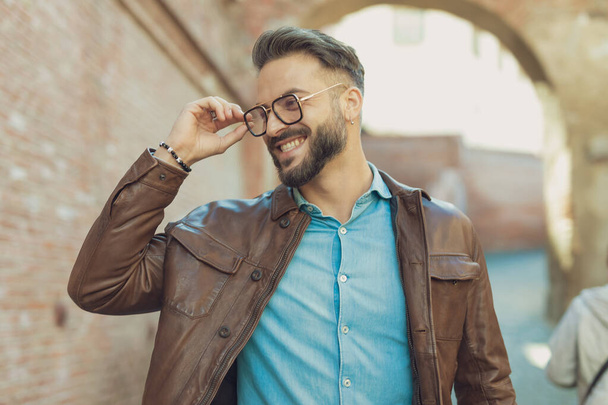 Portrait of attractive casual man arranging his glasses and laughing, wearing leather jacket, outdoor in an old medieval town - Фото, зображення