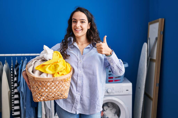 Young brunette woman holding laundry basket smiling happy and positive, thumb up doing excellent and approval sign  - Фото, изображение