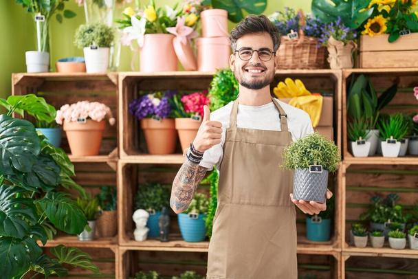 Mladý hispánec s tetováním pracuje v květinářství drží rostlina s úsměvem šťastný a pozitivní, palec nahoru dělá vynikající a schvalovací znamení  - Fotografie, Obrázek