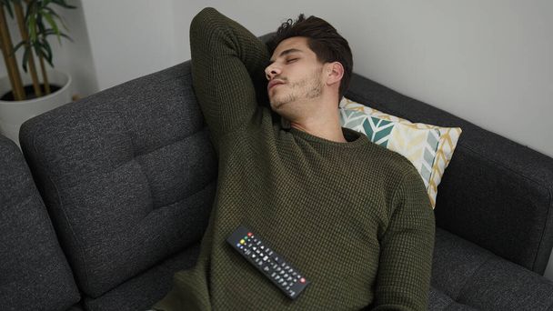 Young hispanic man holding tv remote control lying on sofa sleeping at home - Foto, imagen