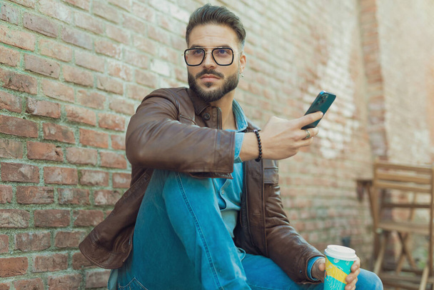Portrait of sexy casual man looking away with a phone and drink, wearing eyeglasses and leather jacket, outdoor in an old medieval town - Fotoğraf, Görsel