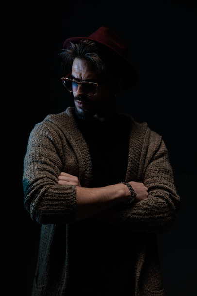 Ritratto di sexy modello maschio che incrocia le braccia e distoglie lo sguardo, indossando un cappello bordeaux, occhiali e cappotto di lana su sfondo scuro da studio - Foto, immagini
