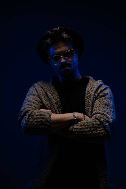 Portrait of attractive casual guy with powerful presence crossing his arms, wearing a burgundy hat, eyeglasses and wool coat in dark studio background - Foto, Imagem