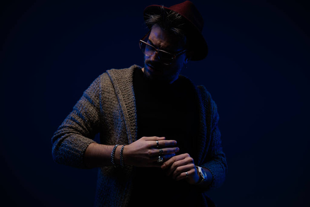 Портрет молодой модели-мужчины с драматической атмосферой, закрепляющей его кольцо, в бордовой шляпе, очках и шерстяном пальто на темном фоне студии - Фото, изображение