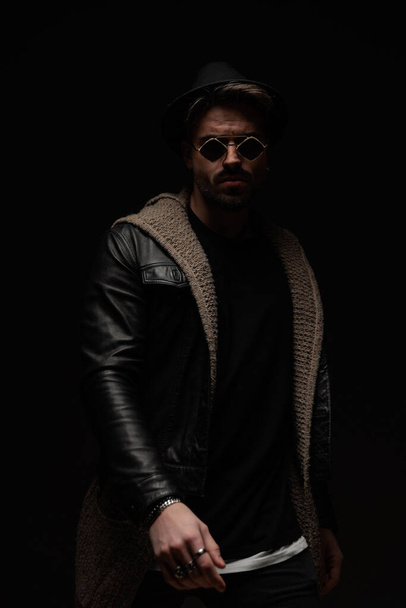 confident bearded man with hat and glasses wearing leather jacket and wool cardigan and posing in a mysterious way on black background - Фото, зображення