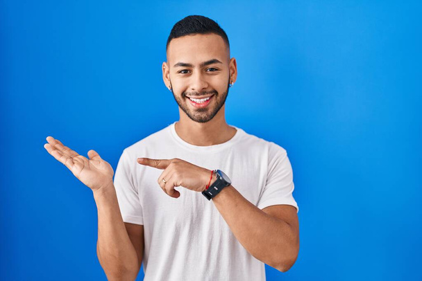 Νεαρός Ισπανόφωνος στέκεται πάνω από το μπλε φόντο έκπληκτος και χαμογελώντας στην κάμερα, ενώ παρουσιάζει με το χέρι και δείχνοντας με το δάχτυλο.  - Φωτογραφία, εικόνα