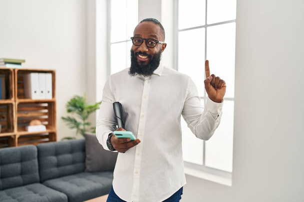 Hombre afroamericano que trabaja en la oficina de terapia usando un teléfono inteligente sonriendo con una idea o pregunta apuntando con el dedo con la cara feliz, número uno  - Foto, imagen