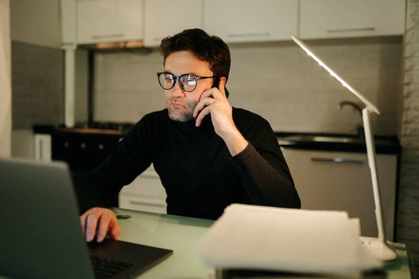 A serious-looking young adult talking on the phone with glasses typing away at his laptop, lost in thought as he works on a project late into the night. - Foto, immagini