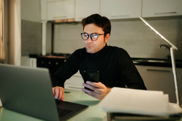 Пізно ввечері онлайн покупки Красивий хлопець в окулярах сидить у своєму ноутбуці, переглядаючи веб-сайти електронної комерції для ідеальної покупки, все з комфорту свого власного будинку
. - Фото, зображення