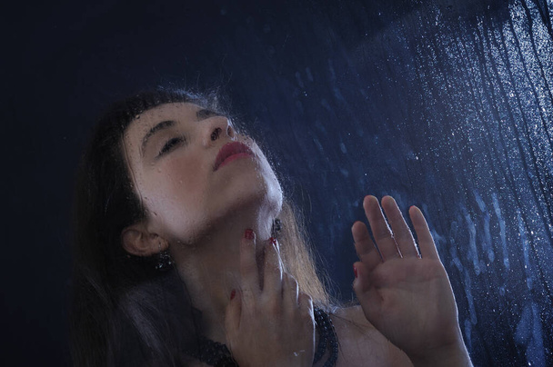 portrait of a beautiful young woman with wet hair and a black face behind window in drop on a dark background - Photo, Image