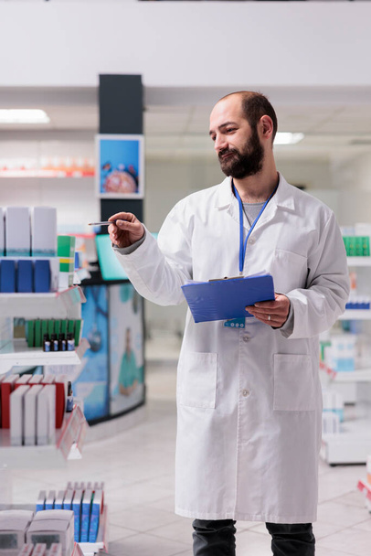 Drugstore employee holding clipboard while checking pills packages writing medication name on papers, working in pharmacy. Pharmacist is responsible for maintaining the inventory of drugs - Zdjęcie, obraz