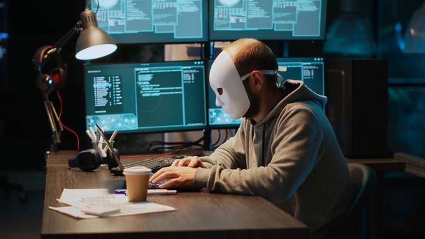 Μασκοφόρος κλέφτης υπολογιστών που χακάρει το σύστημα υπολογιστών αργά το βράδυ, δουλεύοντας σε laptop και πολλαπλές οθόνες για να κλέψει πληροφορίες. Hacker με κρυφή ταυτότητα σπάζοντας διακομιστή ασφαλείας, κυβερνοεπίθεση. - Φωτογραφία, εικόνα