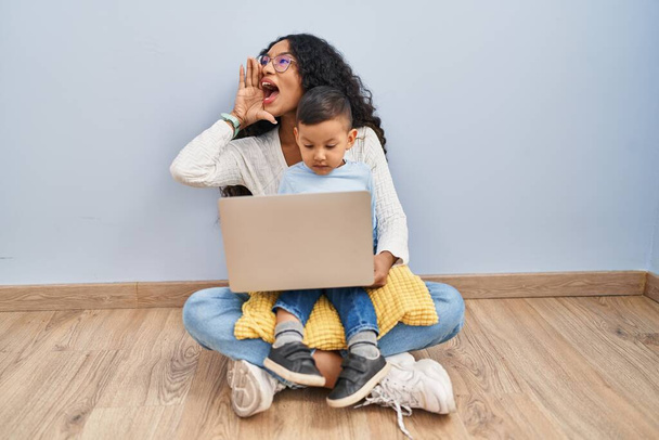 Νεαρή ισπανόφωνη μητέρα και παιδί που χρησιμοποιούν φορητό υπολογιστή κάθονται στο πάτωμα φωνάζοντας και ουρλιάζοντας δυνατά στο πλάι με το χέρι στο στόμα. έννοια επικοινωνίας.  - Φωτογραφία, εικόνα