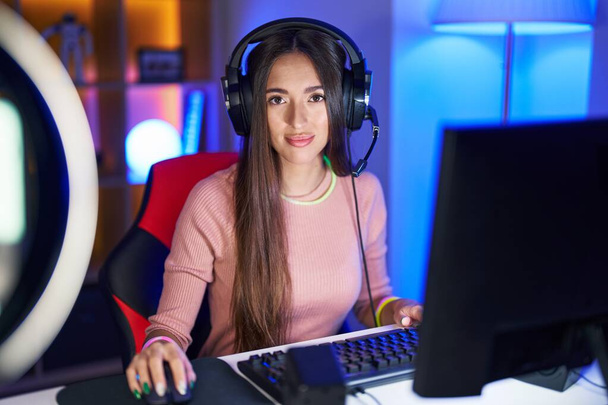 Молода іспаномовна жінка грає в відеоігри розслаблено з серйозним виразом обличчя. простий і природний погляд на камеру
.  - Фото, зображення