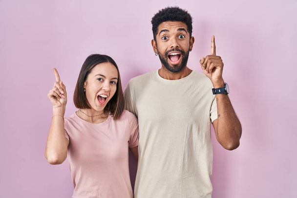 Fiatal spanyol pár együtt rózsaszín háttér mutogatva ujjal felfelé a sikeres ötlet. kilépett és boldog volt. Egyes számú..  - Fotó, kép