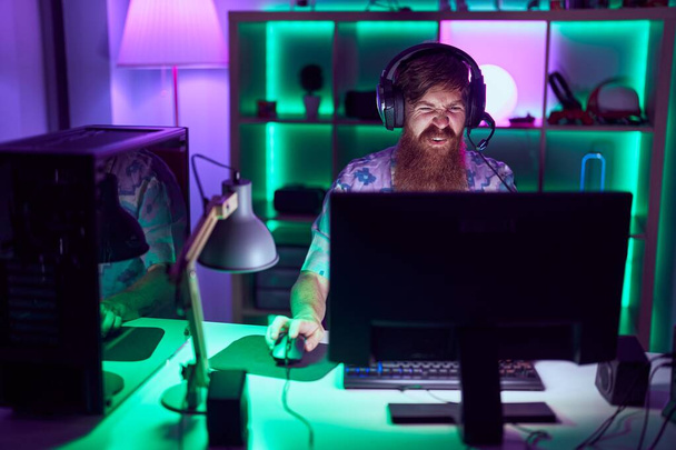 Young redhead man streamer playing video game using computer at gaming room - Photo, Image