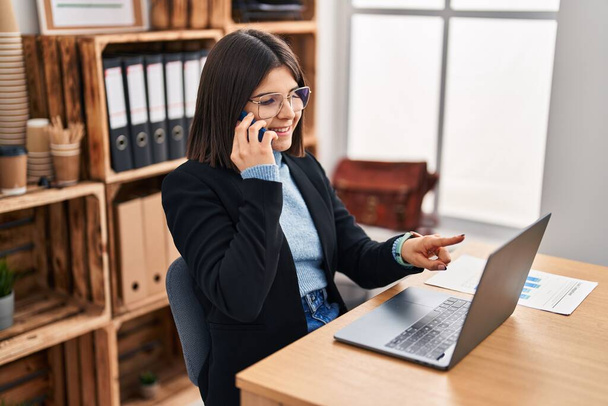 Молода красива іспанська жінка бізнесмен, використовуючи ноутбук розмовляє на смартфоні в офісі
 - Фото, зображення