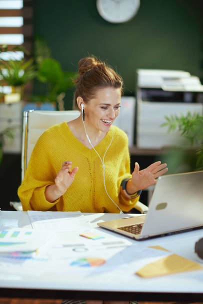 счастливая современная владелица малого бизнеса женщина в желтом свитере с ноутбуком, имеющая веб-камеру встречи в современном зеленом офисе. - Фото, изображение