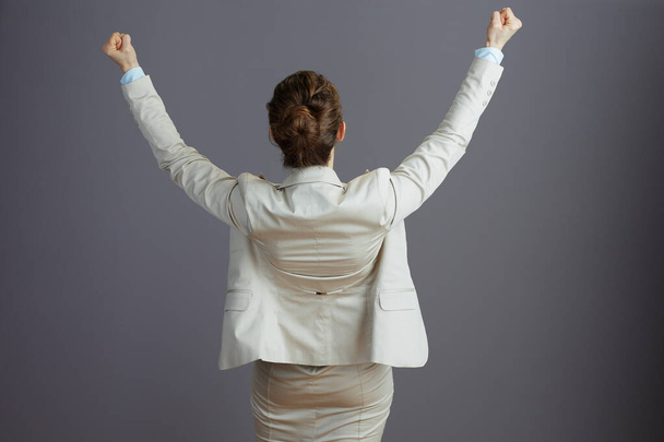 Seen from behind female worker in a light business suit with raised arms rejoicing isolated on gray. - Photo, Image