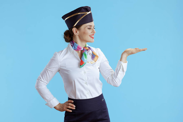 smiling elegant stewardess woman on blue background in uniform presenting something on empty palm. - Photo, image
