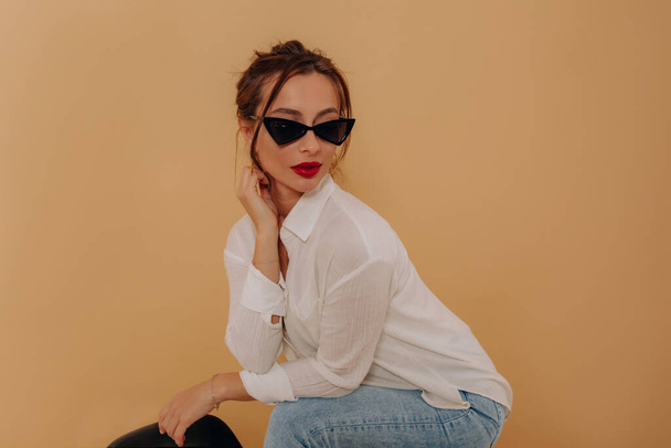 Lovely cute charming girl with dark hair and red lips wearing white shirt, jeans and dark glasses posing to camera during photoshoot over isolated background - Fotografie, Obrázek