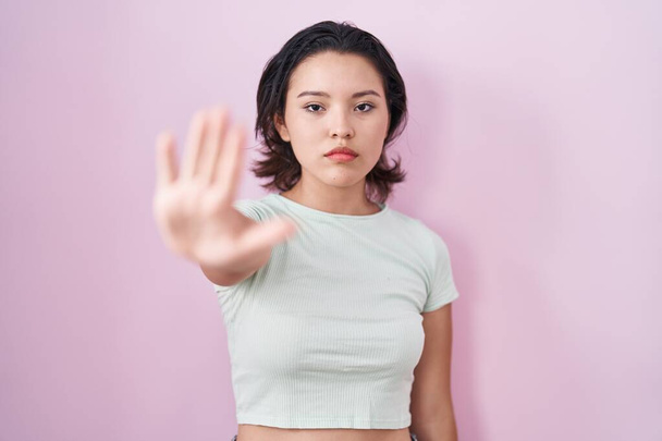 Hispanic young woman standing over pink background doing stop sing with palm of the hand. warning expression with negative and serious gesture on the face.  - Valokuva, kuva