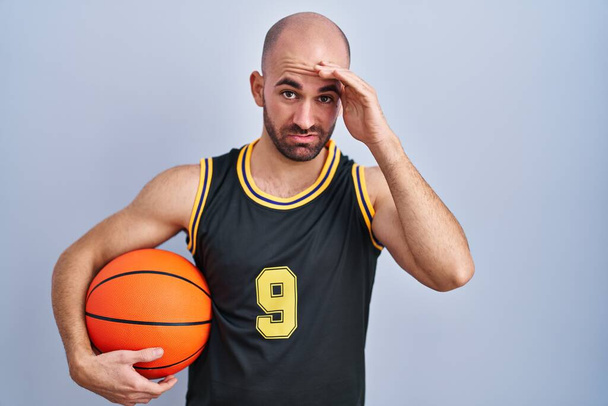 Giovane uomo calvo con la barba indossa l'uniforme da basket tenendo palla preoccupato e stressato per un problema con la mano sulla fronte, nervoso e ansioso di crisi  - Foto, immagini