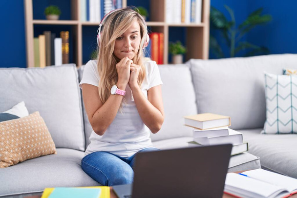 Молодая блондинка учится с помощью компьютера ноутбука дома смеется нервно и взволнованно с руками на подбородке глядя в сторону  - Фото, изображение